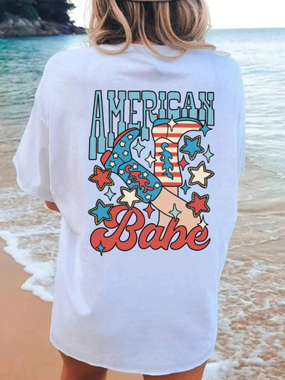 Woman wearing a white beach cover-up with "American Babe" text and star design, standing on a sandy shore with ocean waves in the background.