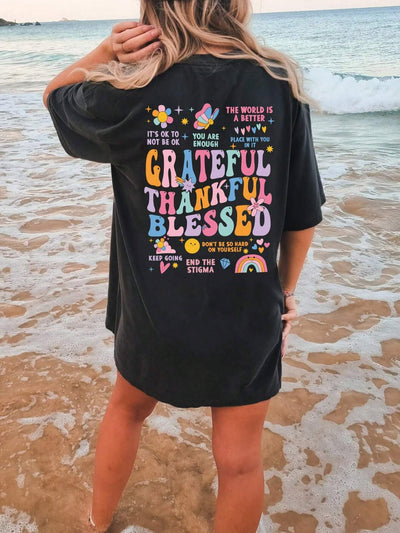 Woman in black t-shirt with colorful motivational quotes stands on sandy beach near ocean waves.