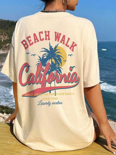 Woman wearing a Beach Walk California graphic T-shirt, sitting by the ocean with palm trees and beach in the background.